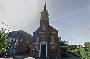 Op de foto is een kerk te zien met daarnaast struiken en een huis. Ook zie je een lantaarnpaal.