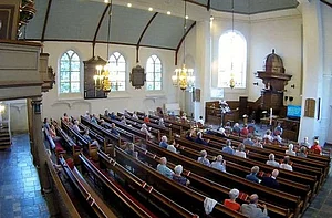 Op de foto zie je meerdere mensen in kerkbanken zitten. Verder zijn ramen, muren en een orgel te zien.