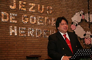 Op de foto spreekt een man met een pak en rode stropdas achter een microfoon. Op de achtergrond staat de tekst 'Jezus de goede herder' op de muur.